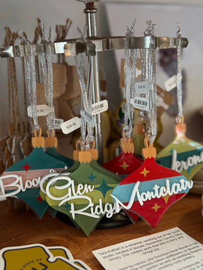 A close-up of several colorful Christmas ornament tags hanging from a silver display stand. The tags are shaped like ornaments and feature names such as Blooming, Glen Ridge, and Montclair in white script, with glittery silver ribbons and small price tags attached.